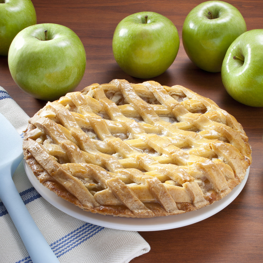 A lattice-crust apple pie on a white plate, with whole green apples arranged around it on a wooden table and a blue spatula at the side.